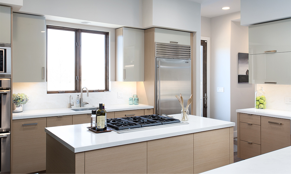 A close-up view of an L-shaped kitchen with white-toned wood veneer cabinets and a central island. The light wood texture adds a natural touch, while the island offers additional workspace and modern style. The partial image highlights the fine craftsmanship and elegant details of the contemporary design.