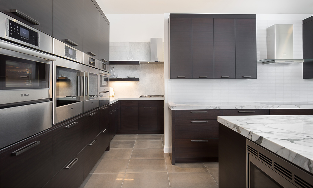 An L-shaped kitchen with dark wood veneer cabinets and a central island, creating a sophisticated and contemporary space. The dark wood finish on the cabinets contrasts beautifully with the sleek island, offering both functionality and a warm, elegant aesthetic.